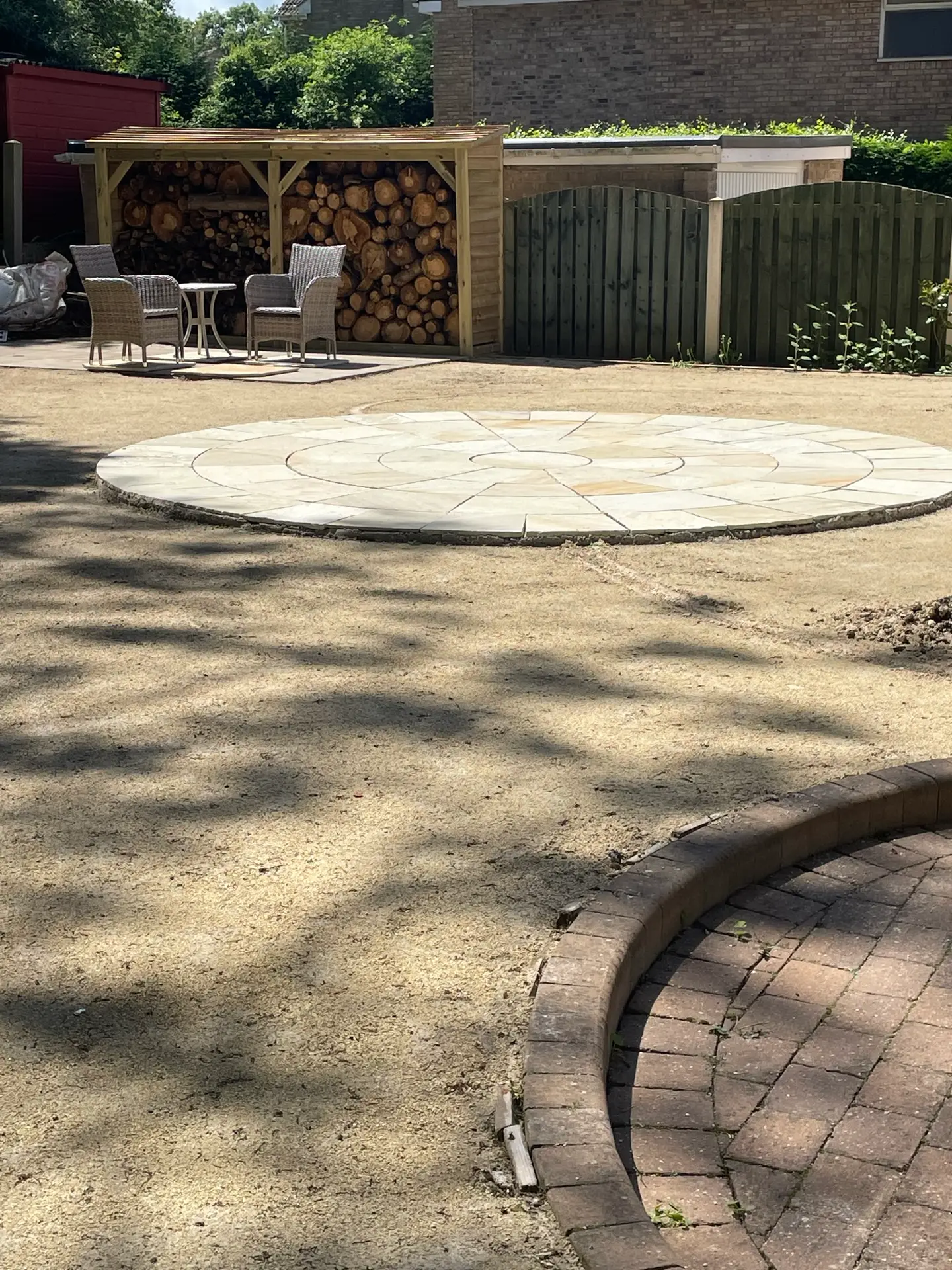 Circular sandstone patio newly laid on bare ground beside a timber log store with two rattan chairs