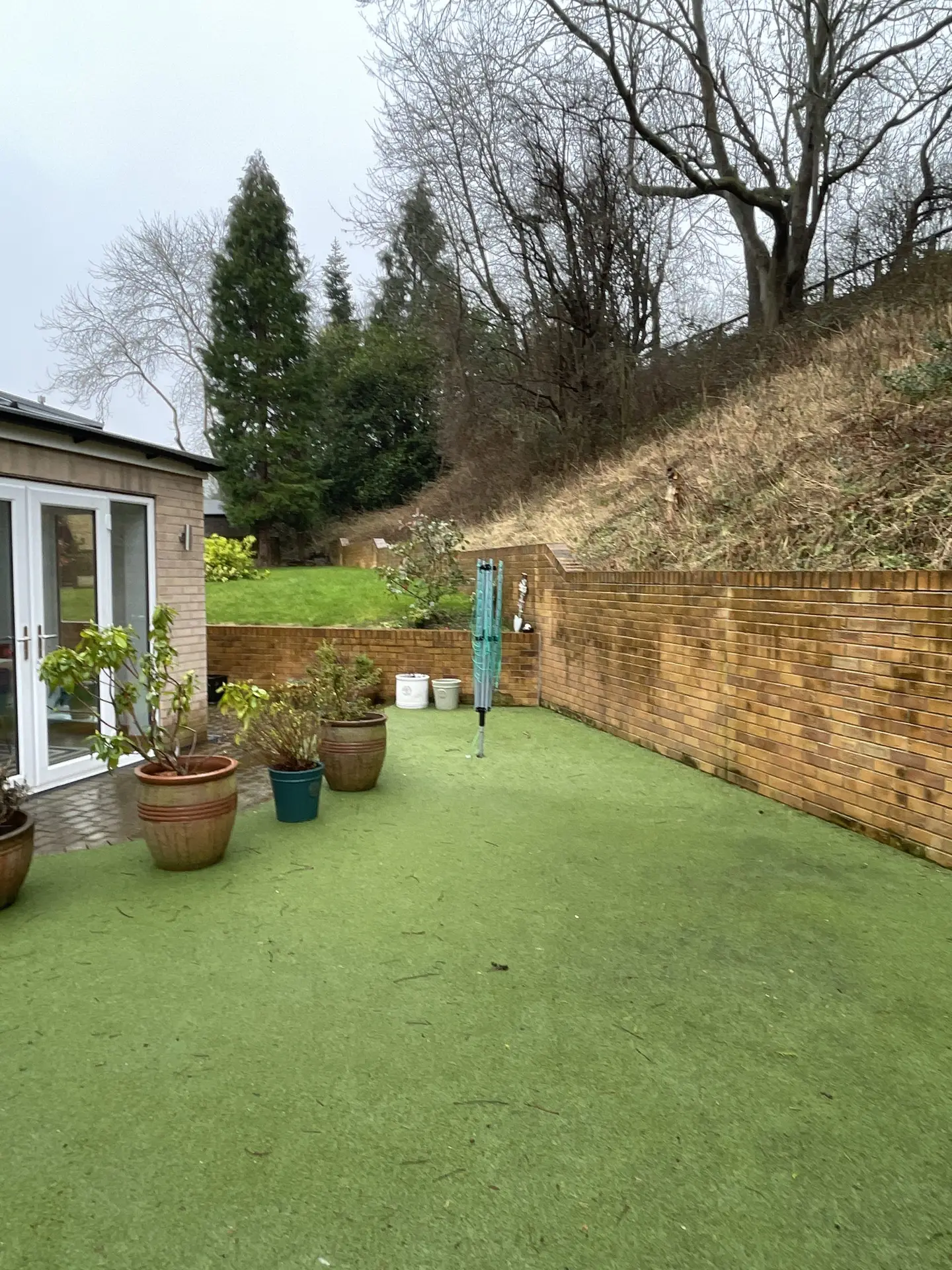 Before: artificial lawn, clothes line and pots below a brick retaining wall and wooded bank