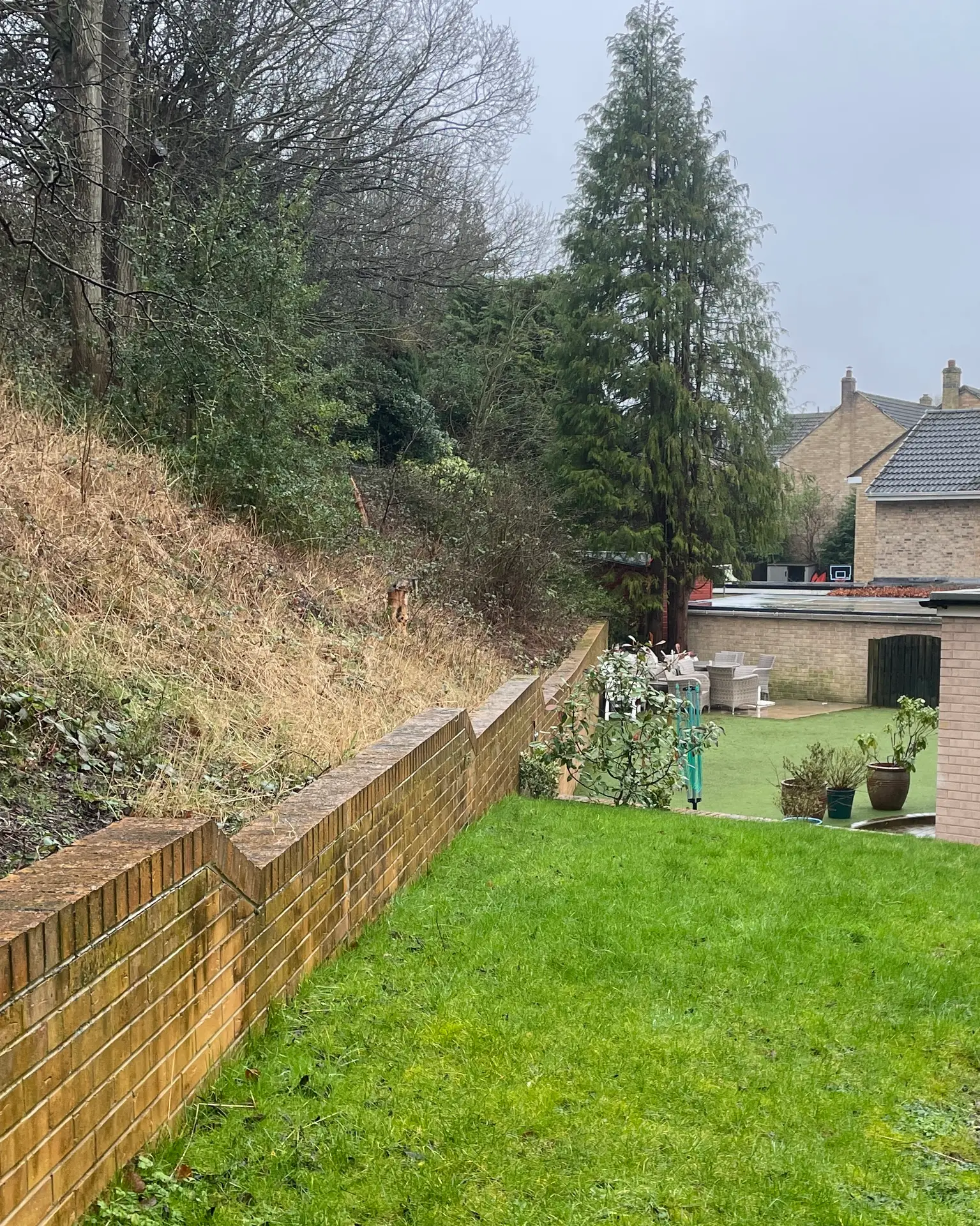 Before: brick retaining wall between a grassy upper lawn and the artificial lawn below, with an overgrown bank above