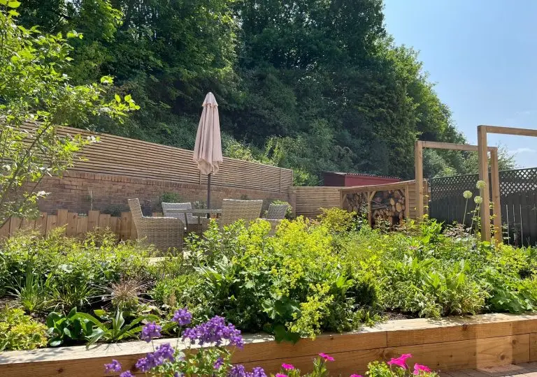 Summer view across a planted border to a patio with umbrella, slatted timber screen, pergola and log store